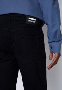 Schwarze Jeans aus Denim mit einem Etikett, auf dem "DRDENIM" steht. Zu den Merkmalen gehören eine glatte Textur, fünf Taschen und eine schmale Passform.