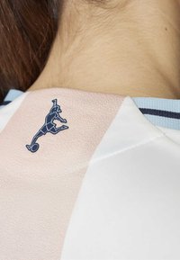 Camiseta deportiva blanca y rosa claro con un emblema azul de una figura saltando con un balón de fútbol en el cuello. Detalle de tejido texturizado visible.
