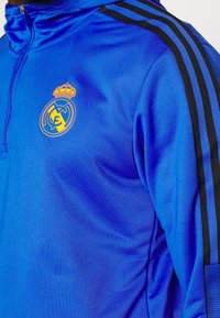 Veste de sport bleue avec des bandes noires sur les épaules, fermeture éclair au milieu du devant, et l'écusson jaune et orange du Real Madrid sur la poitrine.