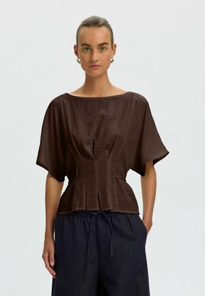 Modèle portant un chemisier marron foncé à manches courtes, cintré à la taille, et un pantalon foncé, devant un arrière-plan uni.