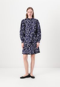 Blumendruck-Kleid in Dunkelblau und Weiß, mit gezacktem Saum, langen Puffärmeln und einer gebundenen Taille. Getragen mit schwarzen Ballerinas.