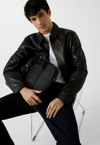 Zwarte leren tas met een gestructureerde vorm, dubbele handvatten, ritssluiting en een logo-accent. Model draagt een zwarte leren jas en donkere spijkerbroek.