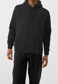 Haut à capuche noir en tissu doux, doté d'une poche kangourou à l'avant, d'une capuche ajustable avec cordon de serrage et de poignets côtelés. Associé à un pantalon en denim foncé.