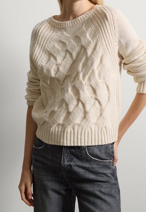 Femme portant un pull en maille torsadée couleur crème avec manches longues remontées, associée à un jean gris foncé sur fond gris clair.