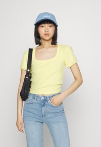 Camisola amarula de manga curta com decote largo, combinada com umas calças de ganga azul claro e um boné azul claro. Bolsa de ombro preta.