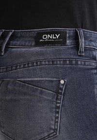 Jeans i denim med mörk tvätt, fem fickor och en svart etikett med varumärket "ONLY". Tyget har en jämn textur och lätt slitage.
