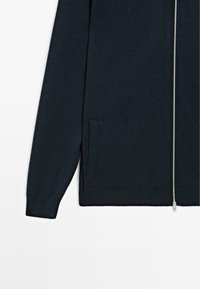 Námo�řnický modrý svetr s plným zipem, dlouhými rukávy a bočními kapsami; vyrobený z jemné látky s hladkou texturou a minimalistickým designem.