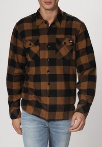 Camicia di flanella marrone e nera con motivo a quadri, dotata di colletto a punta, due tasche sul petto e bottoni neri. Vestibilità classica.