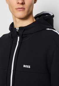 Svart zip-up hoodie med vita accenter, har hög krage och en logotyp, tillverkad av mjukt tyg. Noterbara detaljer inkluderar en dragkedja och sömmar.
