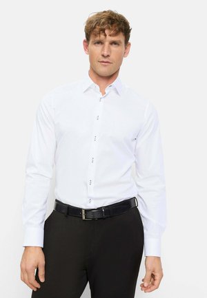 Mann trägt ein eng anliegendes weißes Hemd mit einzigartigen Knöpfen und schwarze Hose, steht vor einem schlichten hellen Hintergrund.