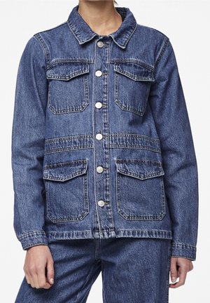 Osoba nosząca niebieską kurtkę denimową z guzikami z przodu i czterema kieszeniami z klapami, w połączeniu z pasującymi spodniami z denimu.