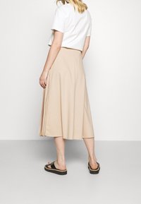 Mujer vestida con una blusa blanca de manga corta, una falda midi beige con pliegues a un lado, y sandalias negras de tipo deslizante, de pie sobre un suelo gris.