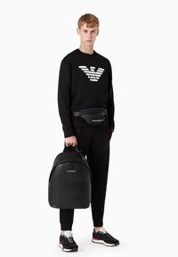 Zaino nero con forma arrotondata e texture liscia, dotato di una tasca frontale e un logo. Indossato da una persona in abbigliamento completamente nero.