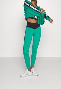 Leggings athletic turchesi con una cintura nera, caratterizzati da strisce laterali. Abbinati a una giacca cropped coordinata e scarpe da ginnastica bianche.