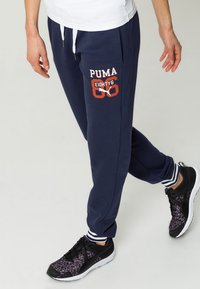 Navy blue joggers with red and white "PUMA EIGHTY6" logo, elastic waistband, and white striped ankle cuffs, paired with black athletic shoes.