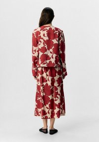 Blomsterprint kjole med lange ærmer, prydet med røde blomster på en beige baggrund. Løs pasform, ankel-længde nederdel og sorte flade sko.