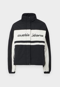 Fekete-fehér puffernadrág magas gallérral, vízszintes csíkokkal és "Guess Jeans" logóval a mellkason, rugalmas mandzsettákkal és elülső zsebekkel.