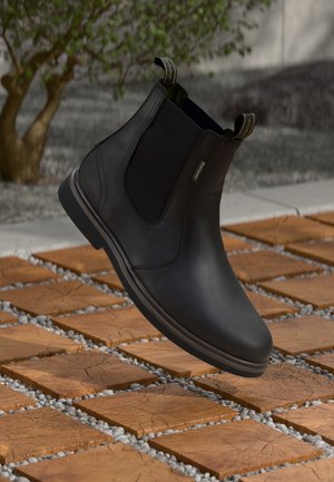 Botas Chelsea pretas em couro com painéis elásticos laterais e alças flutuando sobre azulejos de cubo de madeira e cascalho ao ar livre.