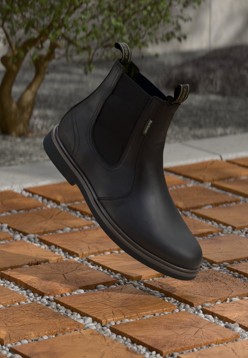 Botas Chelsea pretas em couro com painéis elásticos laterais e alças flutuando sobre azulejos de cubo de madeira e cascalho ao ar livre.