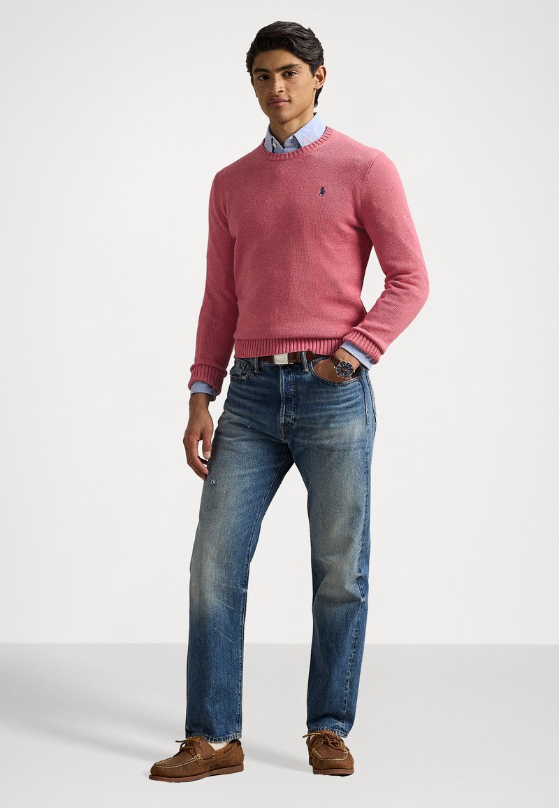 Rosa gestrickter Pullover mit gerippten Bündchen, über einem hellblauen Hemd mit Kragen getragen. Kombiniert mit hellblauen Jeans und braunen Bootsschuhen.