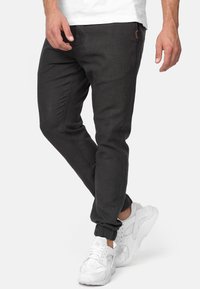 Homme portant un pantalon de jogging noir et des baskets blanches, debout avec une jambe croisée sur l'autre sur un fond blanc.