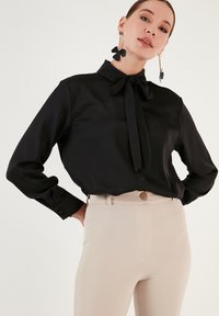 Bluză neagră, cu mâneci lungi, cu un nod la guler, realizată dintr-un material neted, asociată cu pantaloni bej cu talie înaltă, prevăzuți cu un nasture din lemn.