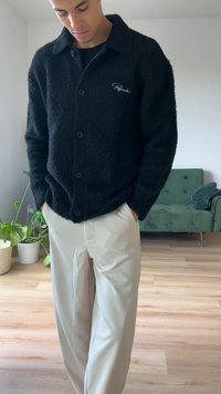 Fekete, bolyhos, gombos dzseki, galléros kialakítással, diszkrét logóval és fekete gombokkal, bézs, bőváló nadrággal viselve.