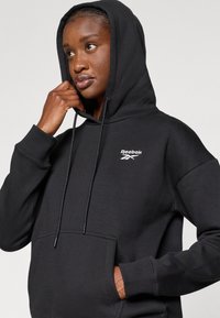Crna dukserica s kapuljačom izrađena od mekog materijala, s prednjim džepom za ruke, rebrastim manžetama i malim bijelim Reebok logom na prsima.