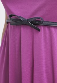 Gros plan d'une ceinture en cuir noir à nœud cintrant la taille d'une robe texturée violette.