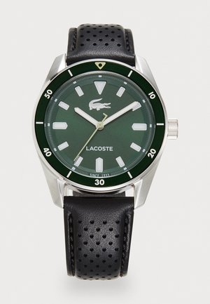 Reloj con carátula verde, caja de acero inoxidable plateada, marcadores de hora blancos, correa de cuero perforado negro y logo de Lacoste en el centro. Incluye detalles de resistencia al agua.