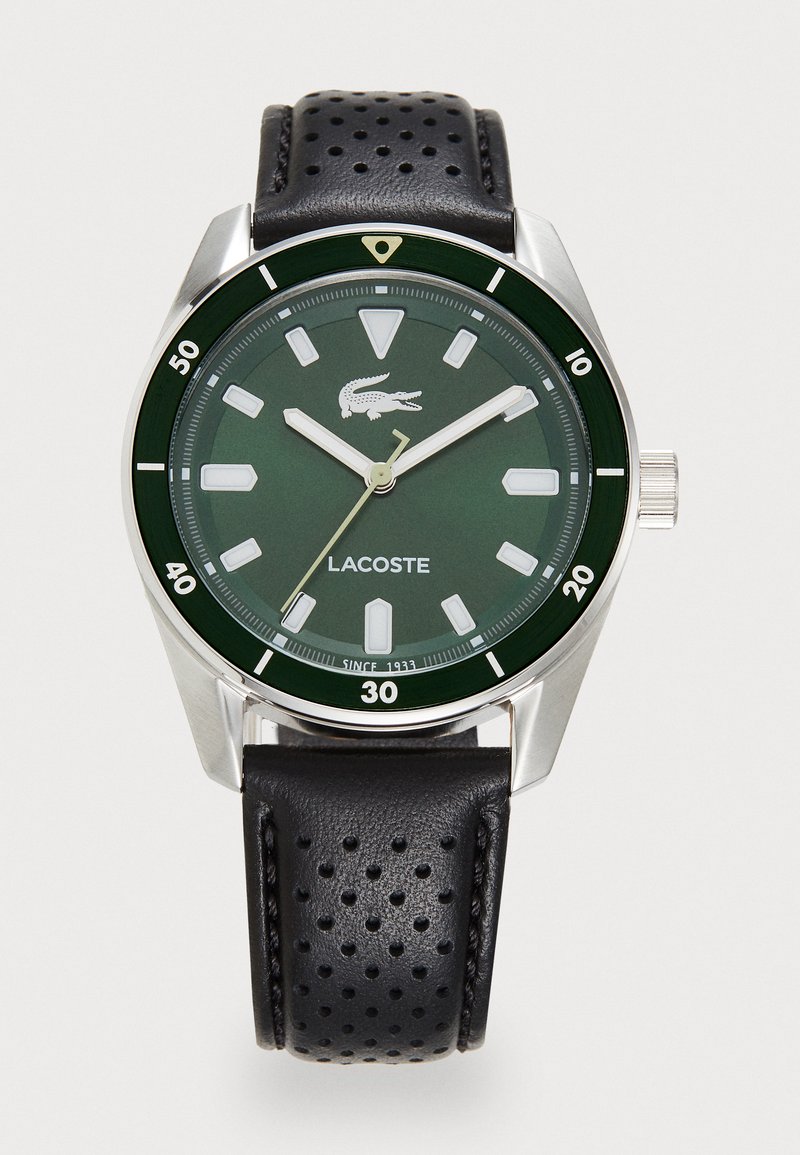 Relógio com mostrador verde e caixa prateada, marcadores de horas brancos, pulseira de couro perfurado preta e logo da Lacoste ao centro. Detalhes de resistência à água incluídos.
