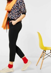 Pantaloni neri su misura indossati con una camicia a fantasia e una sciarpa arancione. Calze rosse, sneakers beige e una sedia gialla sullo sfondo.