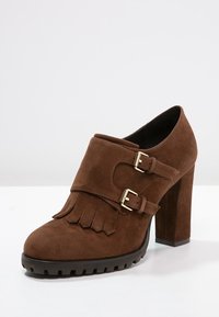 Scarpa alta in suede marrone caratterizzata da dettagli frangiati e due fibbie metalliche regolabili. La suola presenta un design texturizzato e scanalato.