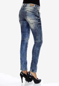Jeans en denim bleu délavé, slim-fit, avec des coutures décoratives, cinq poches et une étiquette en cuir marron au niveau de la taille. Associés avec des talons noirs.