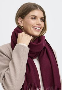 Bordová šatka z mäkk�ého materiálu s ozdobnými strapcami na okrajoch, omotaná okolo krku. Nosená s svetlo béžovým textúrovaným kabátom, vytvára vrstvený vzhľad.