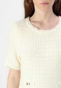 T-shirt en tricot texturé à manches courtes en blanc cassé avec un motif carré. Présente un col brut et un accent en métal décoratif à la taille.
