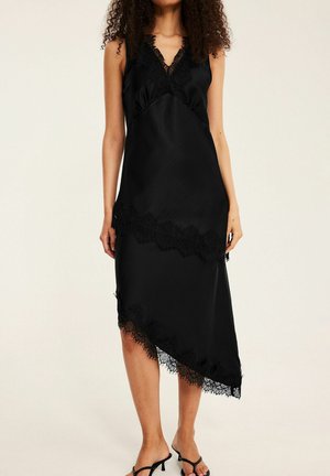 Kvinde iført en sort asymmetrisk midi-kjole uden ærmer med blondekant, stående barfodet mod en ensfarvet baggrund.