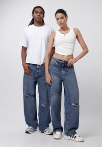 Wide-leg, distressed blue denim jeans with light fading and rips. Styled with a white cropped tank top and a loose white t-shirt.
