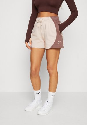 Kobieta w dwukolorowych krótkich spodenkach Nike w beżowo-brązowych odcieniach, białych skarpetkach Nike i białych sneakersach, stojąca z jedną ręką w kieszeni.