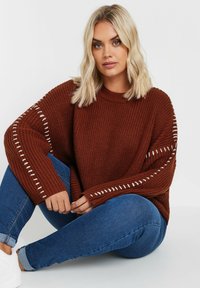 Stickad oversized tröja i rostfärg med kontrasterande cremefärgade sömmar längs ärmarna. Bärs med blå jeans och vita sneakers.