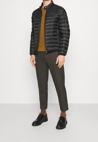 Sort quiltet pufferjakke med høj krave, parret med en brun strikket sweater og skræddersyede brune bukser, båret med sorte snøresko.