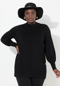 Musta neulepusero, jossa on aaltoileva kaula-aukko ja hihansuut, rento istuvuus sekä kultainen korostusnappi sivulla.