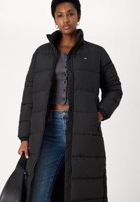 Crni quilted dug puffer kaput s visokim ovratnikom i zatvaranjem na zip. Ima bočne džepove i elastične manžete. Odjeven preko sive košulje s gumbima i trapericama.