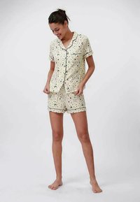 Mujer con conjunto de pijama de manga corta crema con estampado de luna y estrellas, de pie descalza y mirando hacia abajo con una ligera sonrisa.