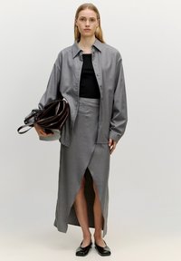 Graues Button-Up-Hemd über einem schwarzen Top, kombiniert mit einem grauen Rock mit hohem Schlitz. Schwarze Schuhe und eine braune Tasche runden das Outfit ab.