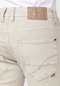 Jeans de mezclilla color beige con un parche de cuero marrón texturizado en la cinturilla. Presenta un simple cosido en los bolsillos traseros y una pequeña etiqueta de tela.