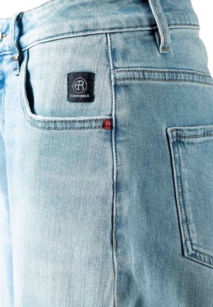 Jasnoniebieskie dżinsy z denimu zbliżenie, pokazujące tylnią kieszeń z czarną metką marki Elias Rumelis oraz detal czerwonego nitu na białym tle.