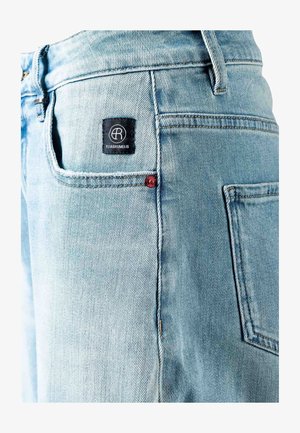 Detailní záběr na světle modré džíny z denimu ukazující zadní kapsu s černou značkou Elias Rumelis a červeným nýtem na bílém pozadí.