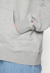 Gray sweatshirt featuring a kangaroo pocket, ribbed cuffs, and hem. Accented with a small red, white, and blue logo on the sleeve.