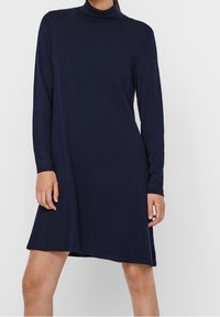 Kvinna i en marinblå långärmad klänning med hög krage som går till knäna och har en rak siluett, mot en neutral bakgrund.